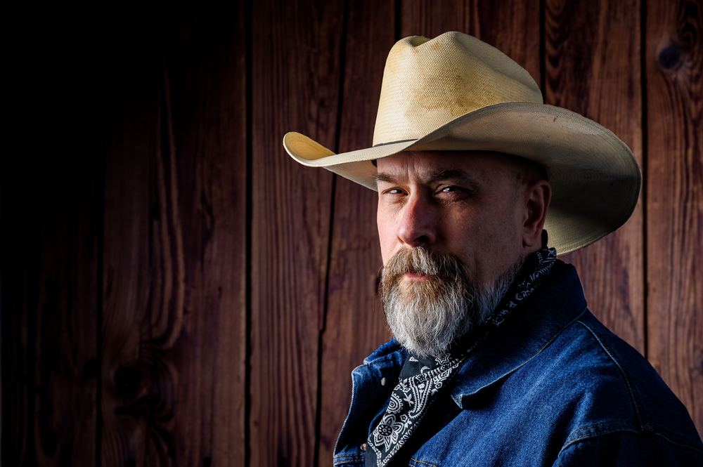 Portrait of a rugged rancher in a straw cowboy hat and denim jacket against a wood barn wall